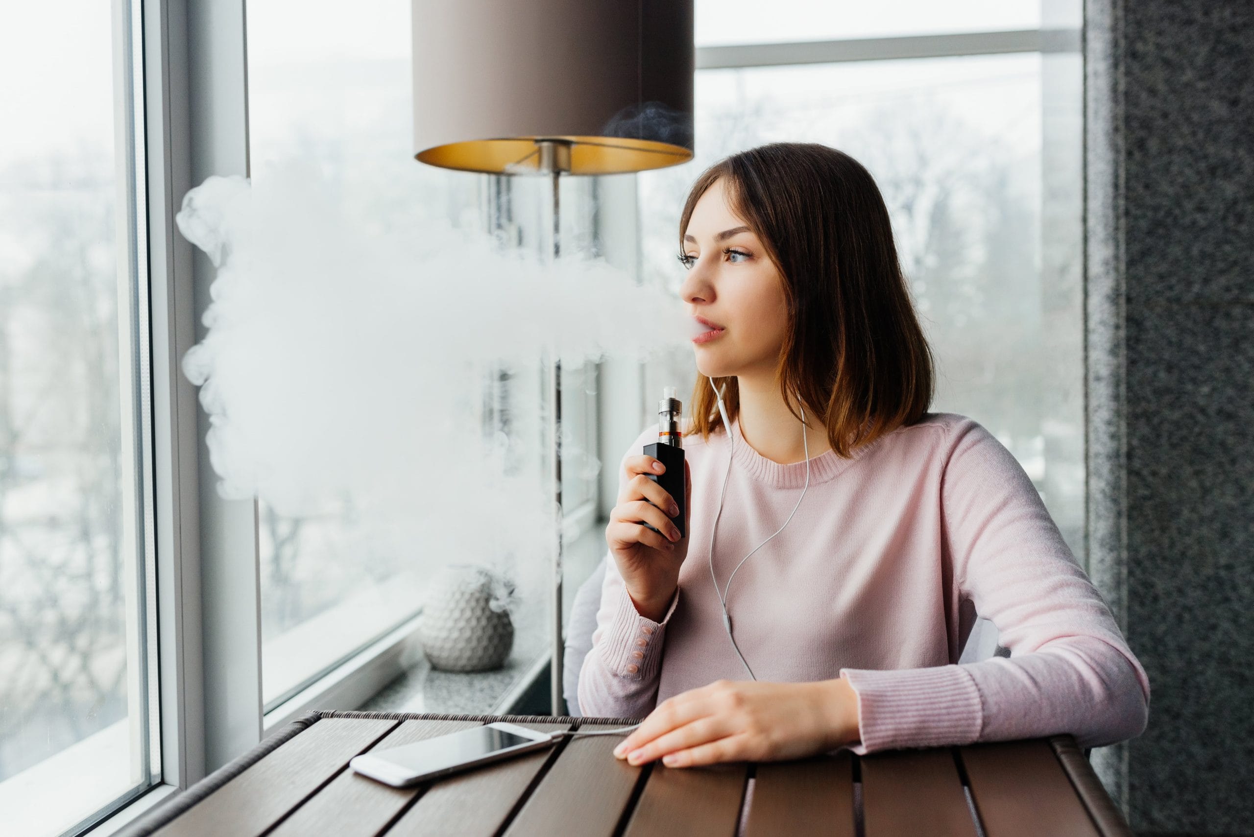Young woman vaping indoors near a window, exhaling thick vapor while holding an electronic cigarette, wearing a pink sweater and earphones.