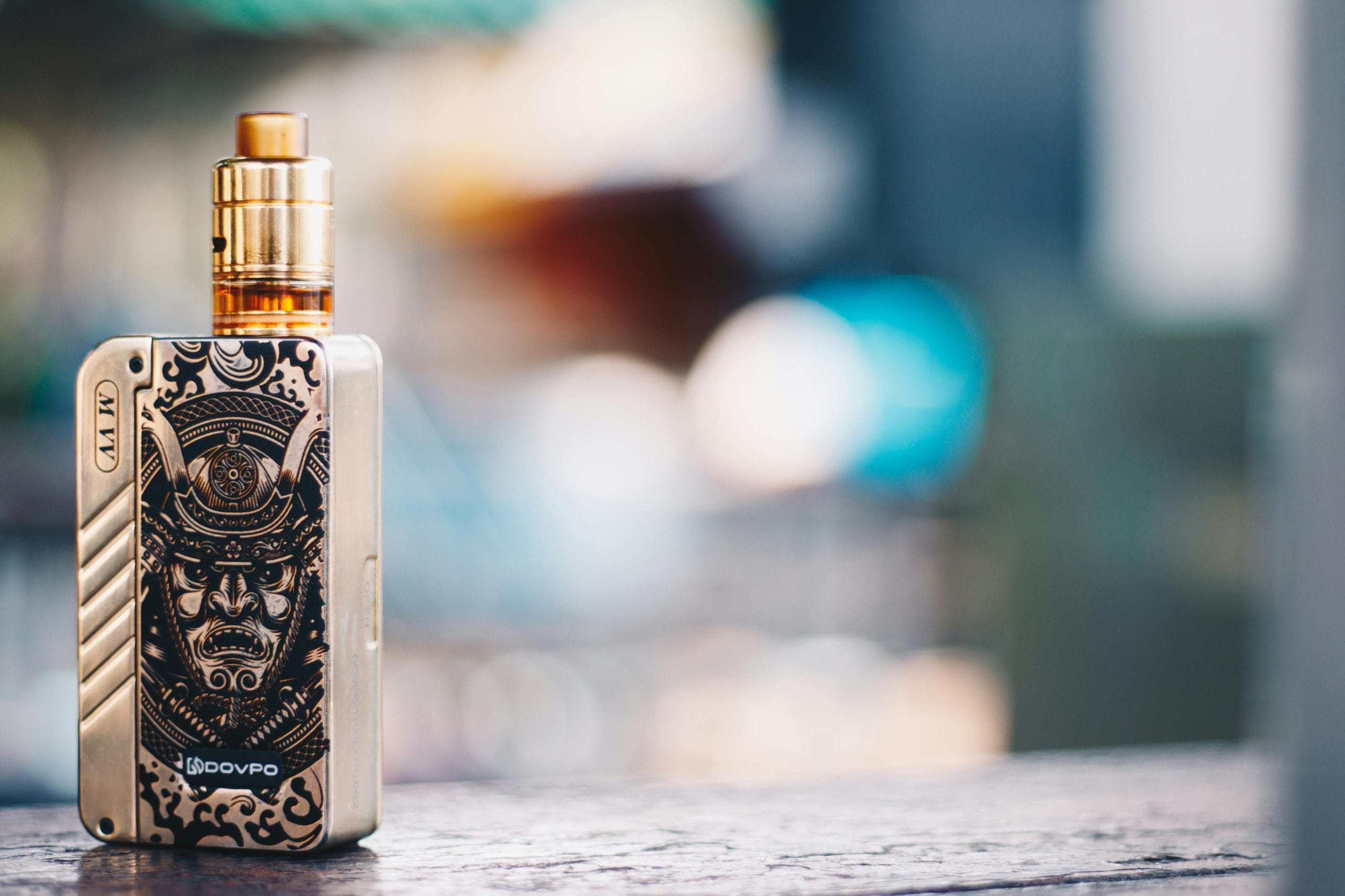 Stylish gold vape mod with intricate samurai design placed on a wooden surface, with a blurred cityscape background.