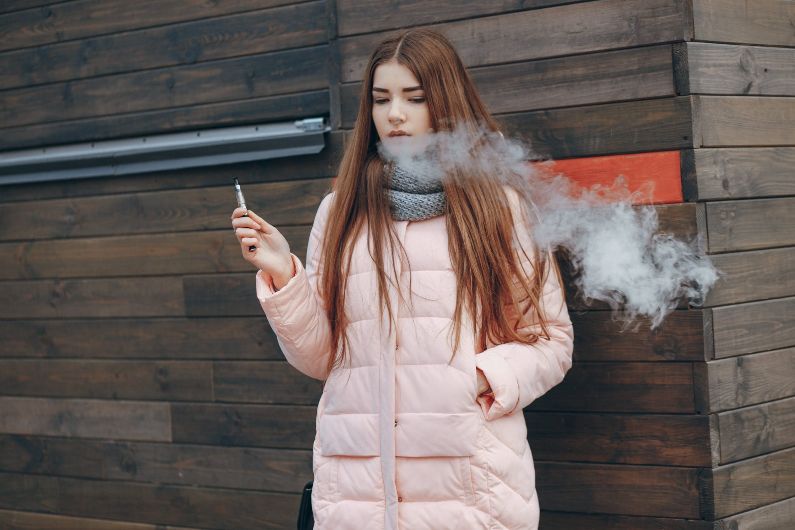 Young woman vaping outdoors in winter coat, exhaling vapor cloud near wooden wall – vape lifestyle concept