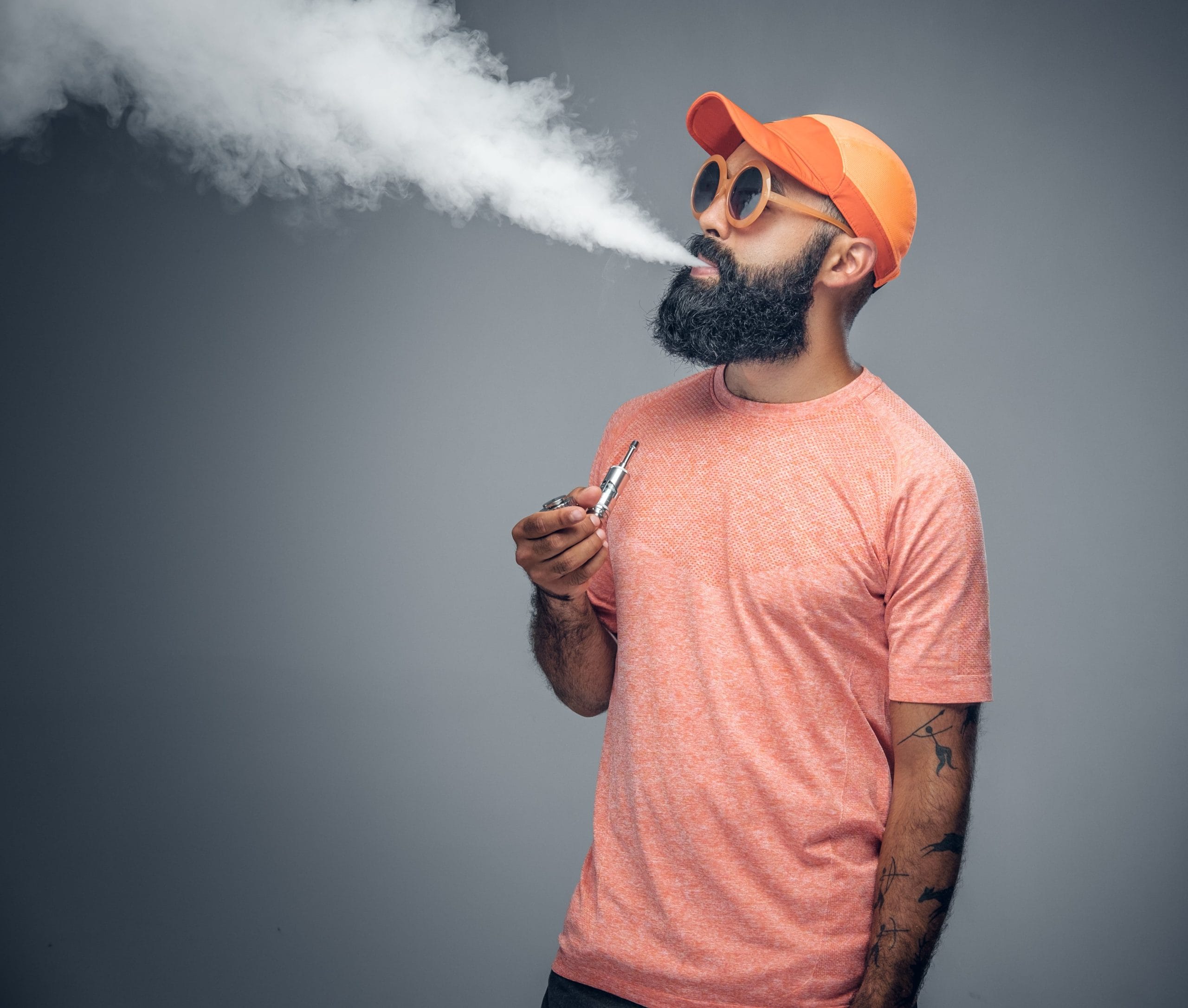 man with cool facial hair wearing pink t-shirt with sunglasses and an orange cap holding a vape and puffing smoke