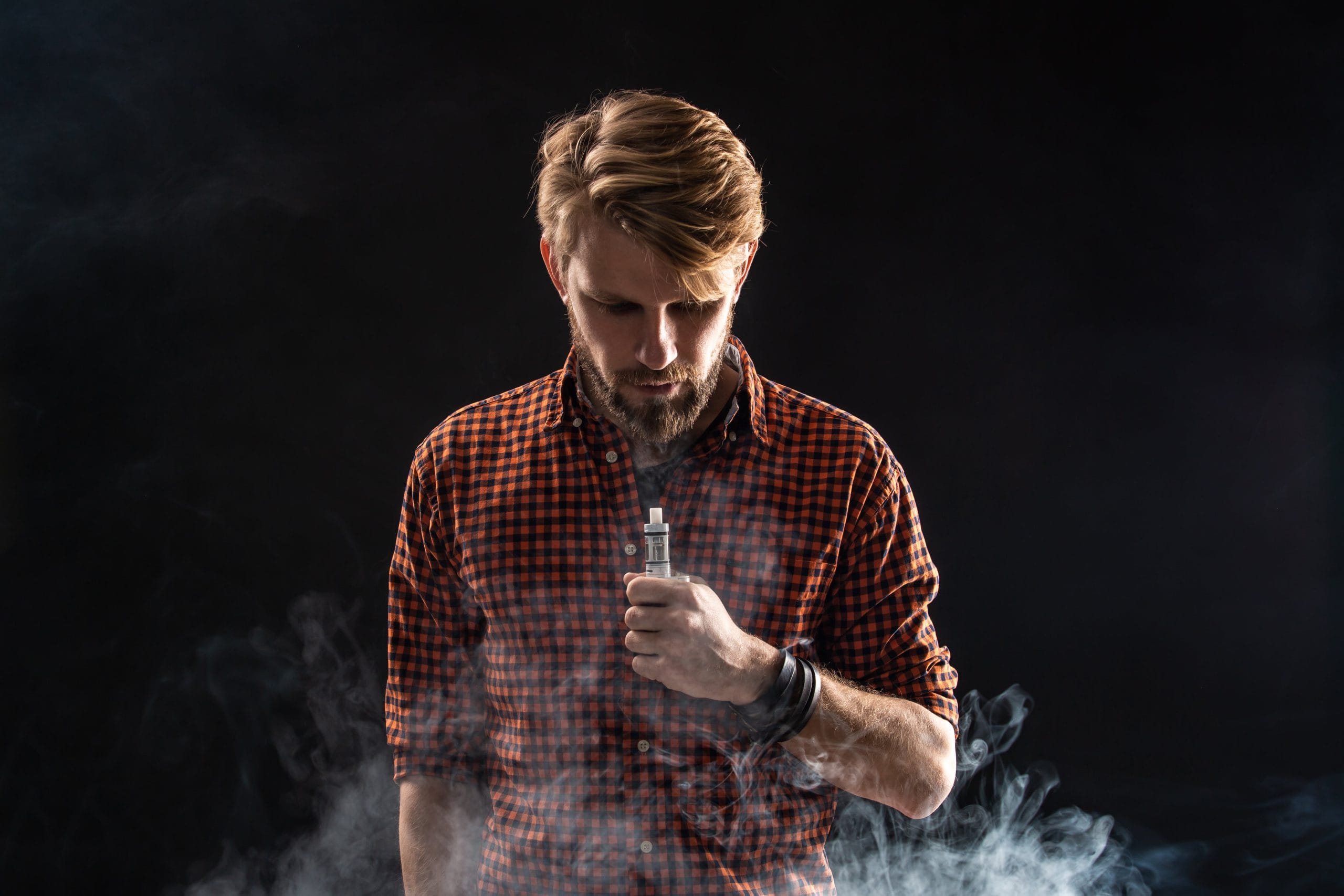 Bearded man vaping in studio with smoke clouds against dark background, wearing checkered shirt – vape lifestyle concept