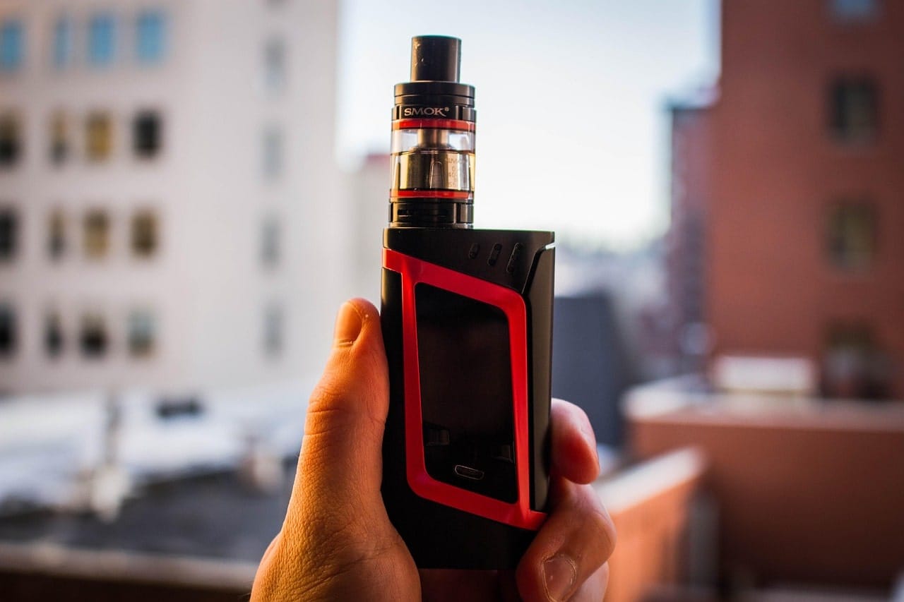 Hand holding a black and red SMOK vape device outdoors in Dallas cityscape background